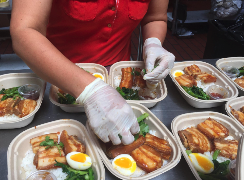 Lish box lunch catering features eco-friendly boxes, with a person in gloves preparing meals of meat, egg, and greens.