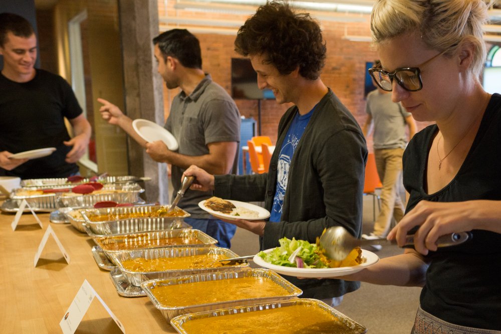 A group of coworkers serves themselves from a Lish buffet with several hot food options in catering trays.