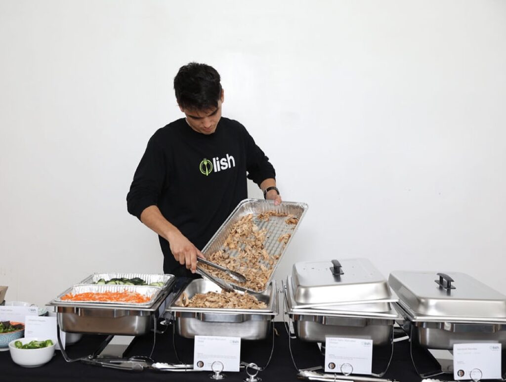 A Lish employee serves food from a catering buffet with a black tablecloth and white walls in the background.