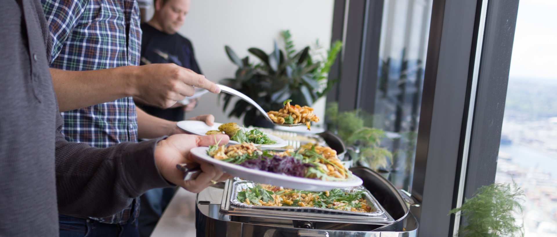 A Lish-catered office lunch buffet with people serving themselves against a backdrop of a high-rise window view.