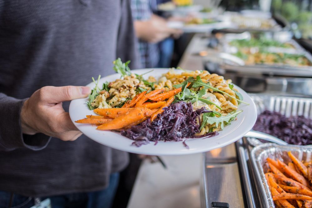 A person holds a plate with food from a Lish lunch catering buffet featuring roasted carrots, pasta, and red cabbage.