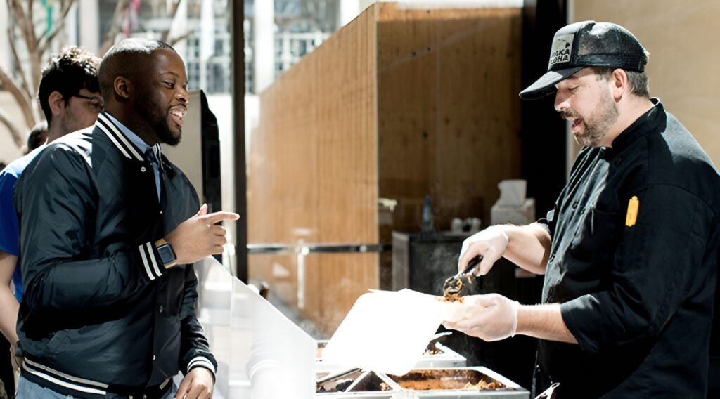 An employee in a jacket is being served food by a Seattle caterer from Lish in a black shirt and baseball cap.