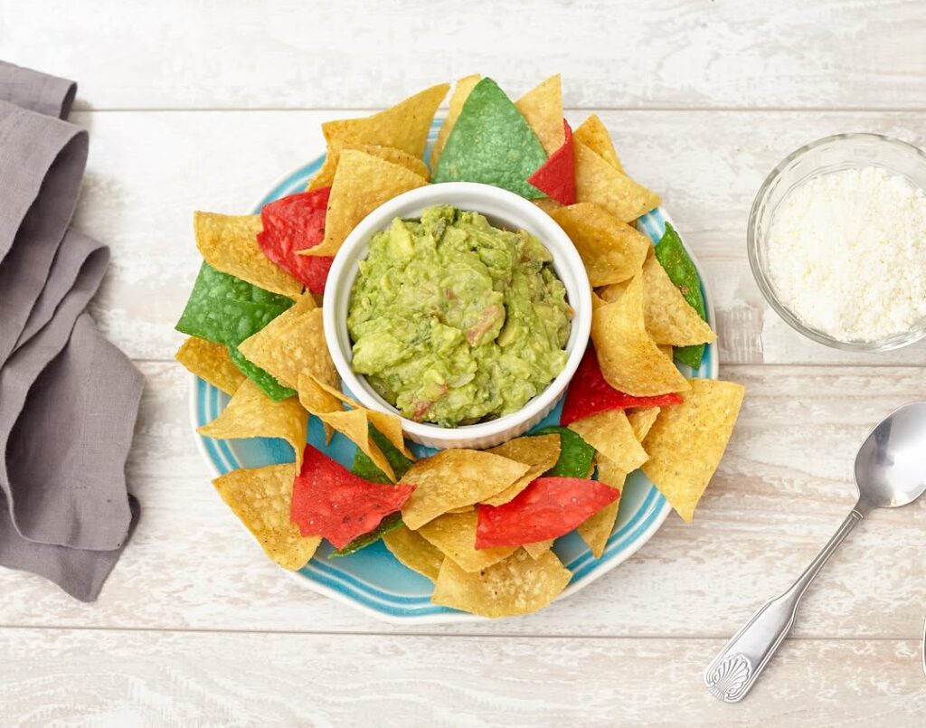 Lish Mexican catering, including Guacamole and colorful tortilla chips on a blue plate, with Cotija cheese on the side.