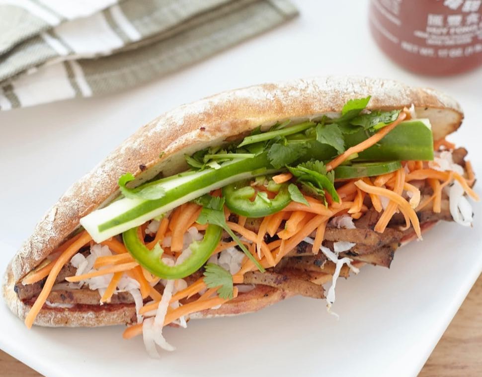 Sandwich from Lish Foods filled with grilled meat, shredded carrots, jalapenos, and cilantro on a white plate.