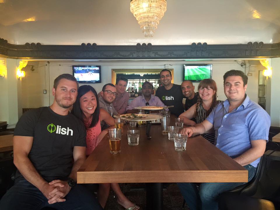A group of Lish employees seated around a large wooden table enjoying drinks and food together.