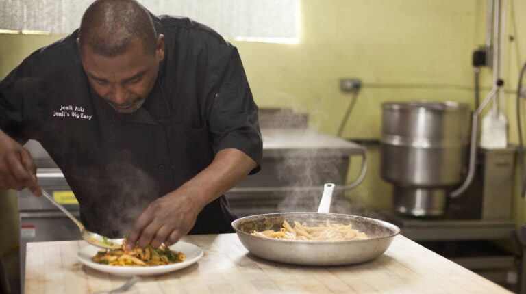 A Lish chef plates steaming pasta from a pan onto a white dish in a commercial kitchen.
