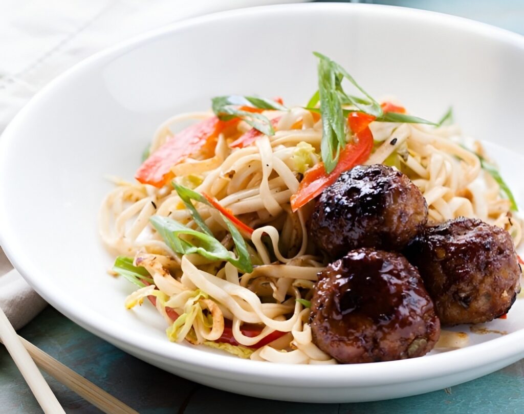 A plate of stir-fried noodles with vegetables, green onions, and meatballs from Lish.