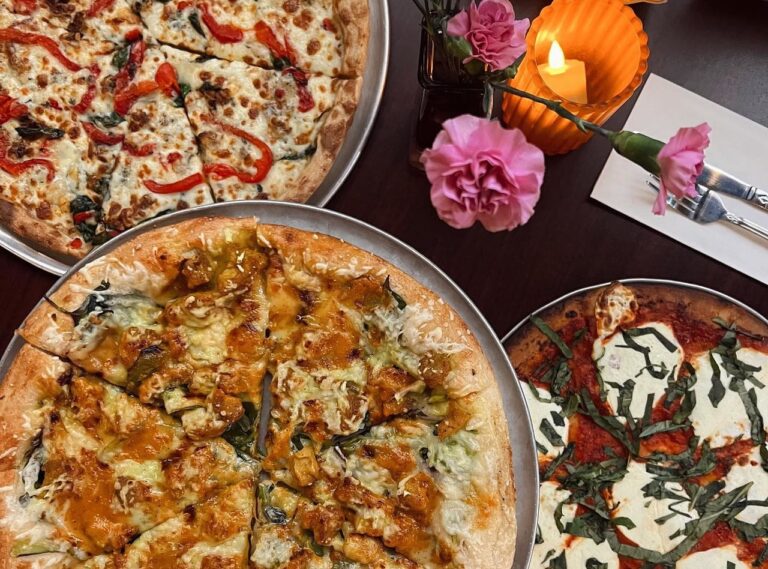 An overhead view of three artisanal pizzas catered by Lish on a dark table, with lit candles and pink flowers.