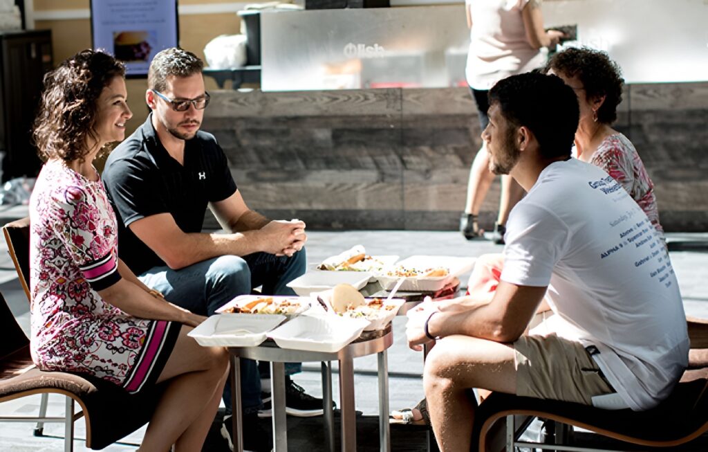 Four employees smiling, talking, and enjoying Individual meal boxes catered by Lish in the office cafeteria.